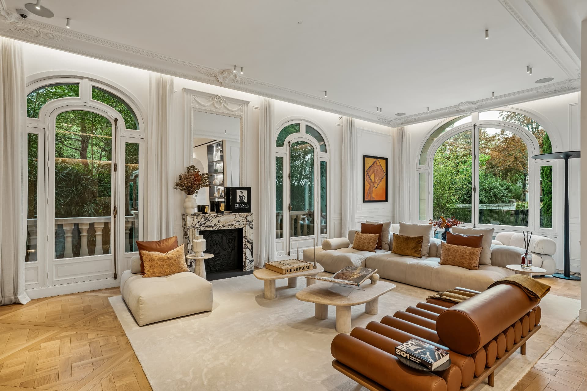 Salon lumineux aux murs blancs moulurés et parquet en chevrons, canapé beige et banquette en cuir cognac, grandes portes-fenêtres ouvertes sur un jardin