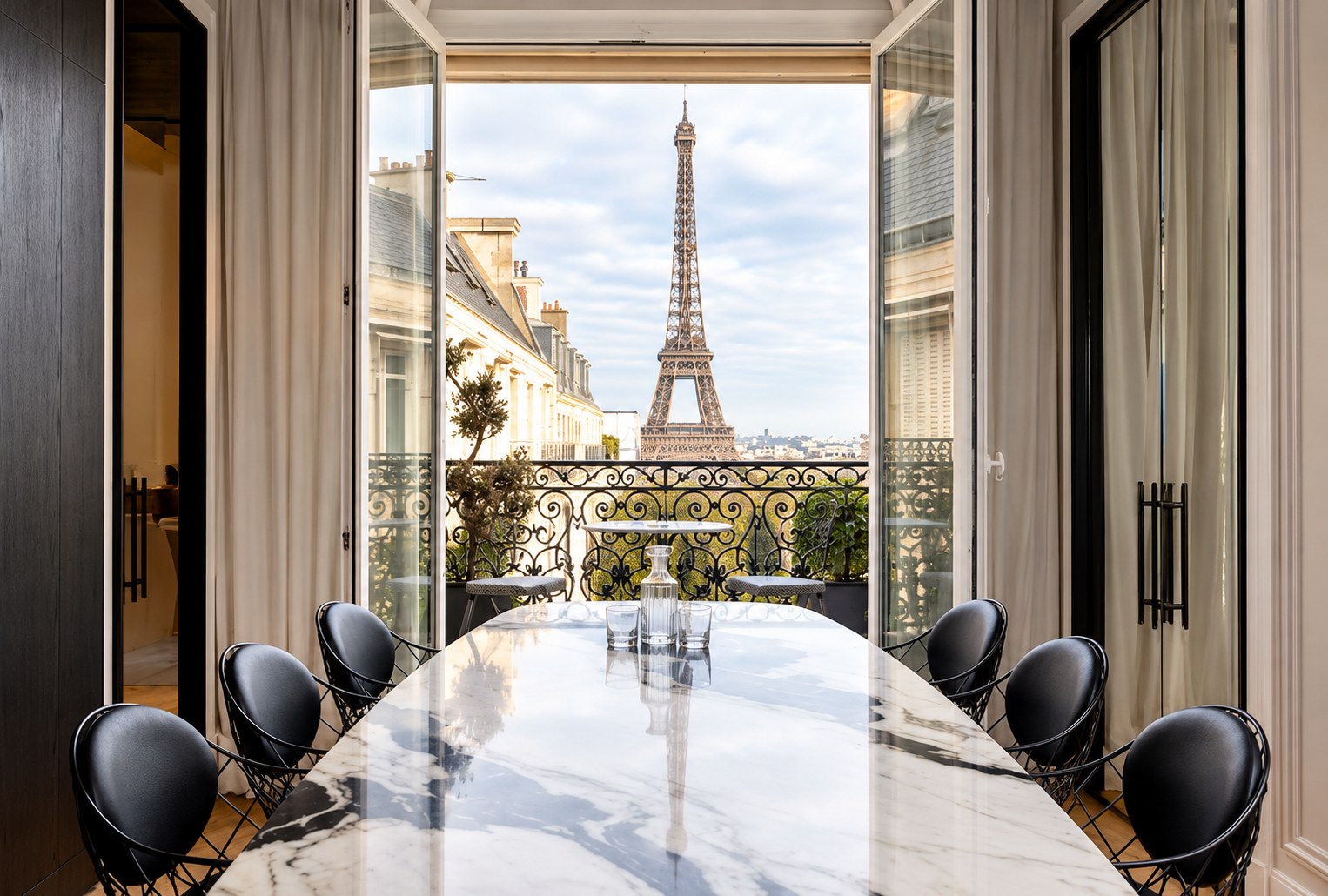 Salle à manger aux chaises noires autour d’une table en marbre blanc, portes-fenêtres ouvertes sur un balcon en fer forgé découvrant la tour Eiffel